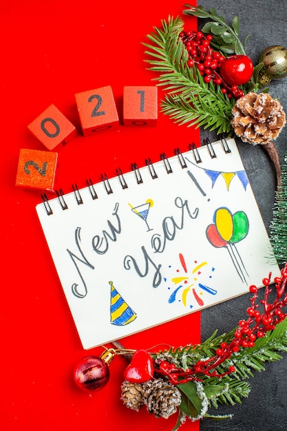 Free Photo vertical view of spiral notebook with new year writing and drawings decoration accessories fir branches xsmas sock numbers on a red napkin and christmas tree on dark background