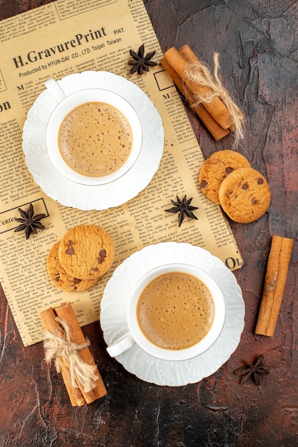 Vertical view of two cups of coffee cookies cinnamon limes on an old newspaper on dark background