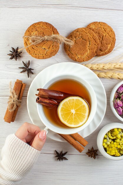 Vertical view of woman hand holding a cup with black tea with cinnamon lime lemon and various herbals cookies on white background