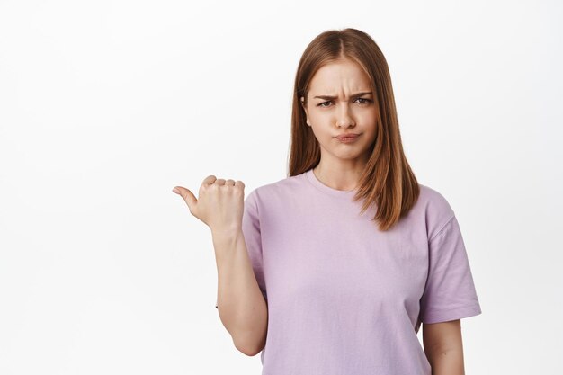 Very bad. Disappointed blond girl complains, grimacing and furrow eyebrows, frowning from dislike, pointing left at smth unpleasant, standing displeased against white background.