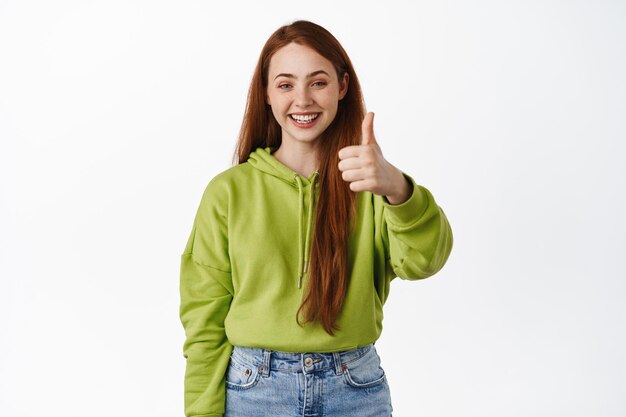 Very good. Smiling redhead teen shows thumbs up and looks satisfied, praise great deal, recommending store, make compliment, white background.