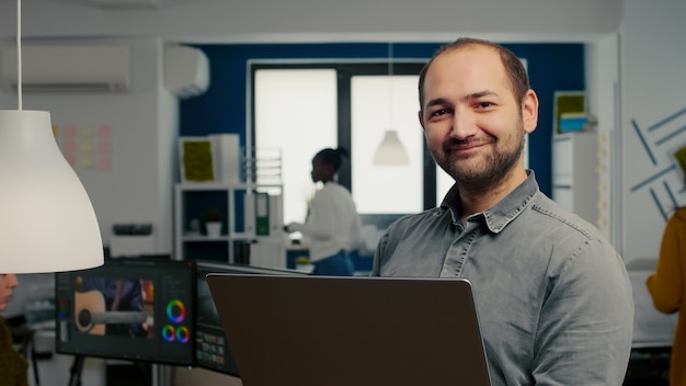 Video editor worker standing in front of camera smiling working in creative agency office holding la...