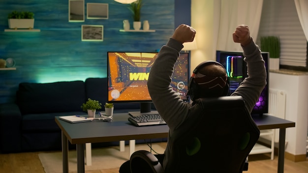 Videogamer player raising hands after winning first person shooter competition wearing hradphones. Professional pro gamer playing online video games with new graphics on powerful computer