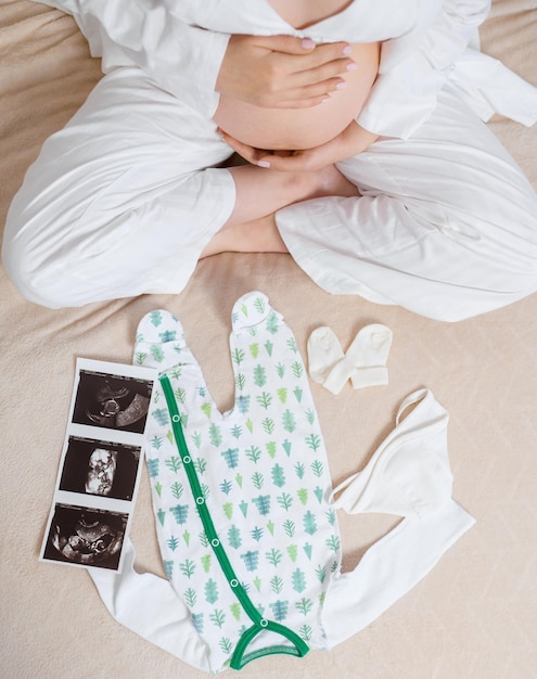 Above view of anonymous woman in white cozy pajama sitting on bed with ultrasound image of womb cute bodysuit and socks while caressing pregnant belly and expecting for baby and part of family