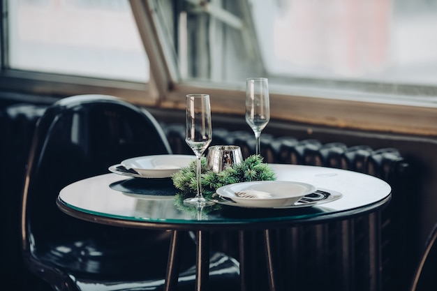 Free Photo view over beautifully decorated round table with natural fir branch, candle, two flutes, plates against classic sofa in modern apartment.