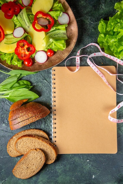 Free photo above view of black bread slices fresh chopped vegetables on a plate and meters green bundle spiral notebook on dark colors surface