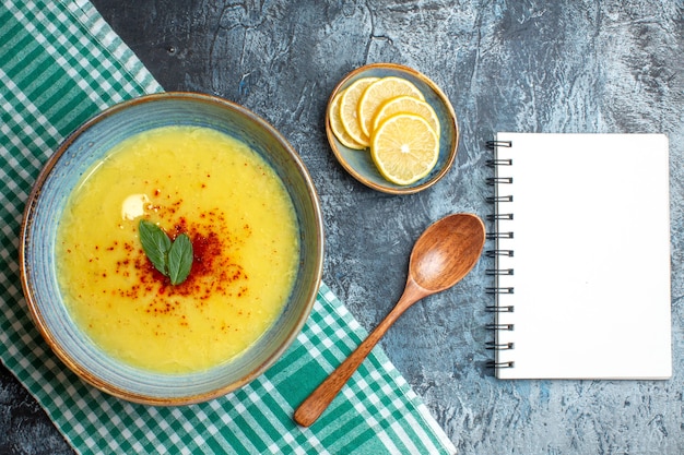 Above view of a blue pot with tasty soup served with mint and pepper next to chopped lemon wooden spoon spiral notebook on blue background