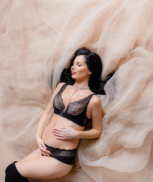 Free Photo above view of charming brunette with closing eyes lying on beige weightless fabric wearing in elegant black panties and bra expecting for baby while touching pregnant belly