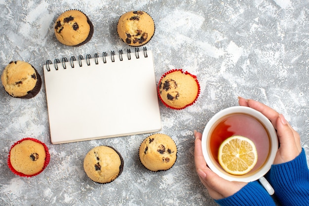 Free photo above view of closed notebook among delicious small cupcakes with chocolate and hand holding a cup of black tea with lemon on ice surface