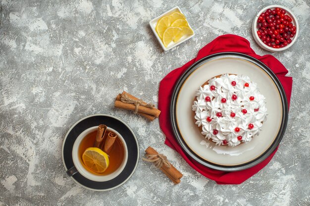 Above view of creamy cake decorated with fruits on a red towel and a cup of black tea with cinnamon limes