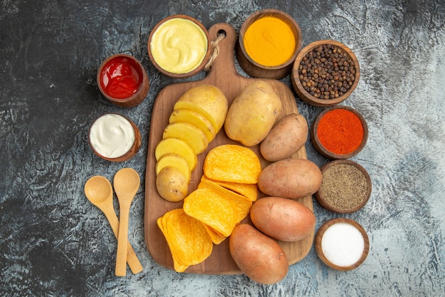 Above view of crispy chips and uncooked potatoes on wooden cutting board and different spices mayonnaise ketchup on gray table