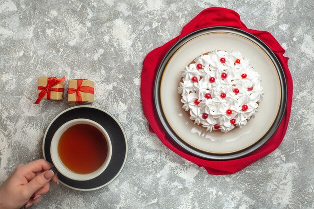 Above view of delicious creamy cake decorated with fruits on a red towel hand holding a cup of black tea small gift boxes on ice background