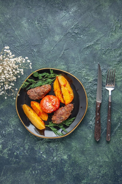 Free photo above view of delicious meat cutlets baked with potatoes and tomatoes on a black plate cutlery set white flowers on green black mixed colors background