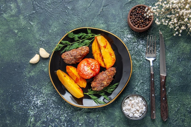 Free photo above view of delicious meat cutlets baked with potatoes and tomatoes on a black plate garlics spices cutlery set on green black mix colors background