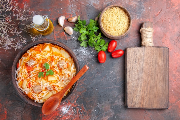 Above view of delicious noodle soup with chicken and uncooked pasta in a small brown bowl and spoon garlic tomatoes and greens cutting board on the dark background