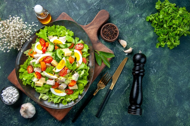 Above view of delicious salad with fresh ingredients on wooden cutting board spices oil bottle garlics cutlery set on black mix colors table