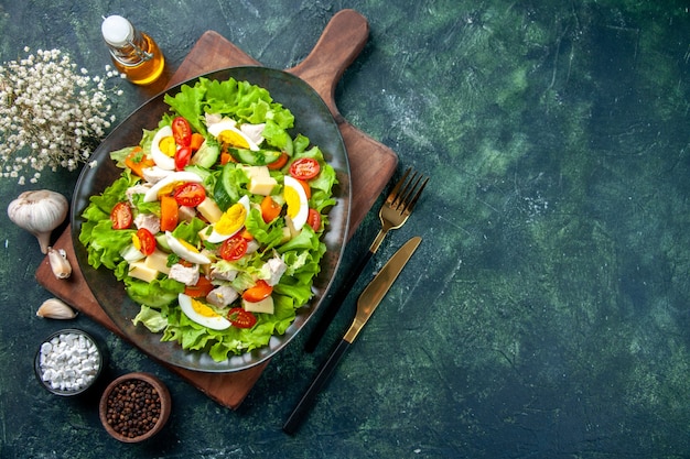 Above view of delicious salad with many fresh ingredients on wooden cutting boa rd spices oil bottle cutlery set on the right side on black green mix colors table