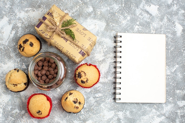 Above view of delicious small cupcakes and chocolate in a glass pot next to Christmas gift and closed notebook on ice surface