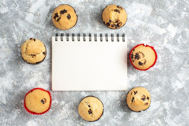 Above view of delicious small cupcakes with chocolate around closed notebook on ice surface