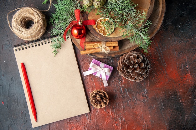 Above view of fir branches and closed spiral notebook with pen cinnamon limes conifer cone and ball of rope on dark background