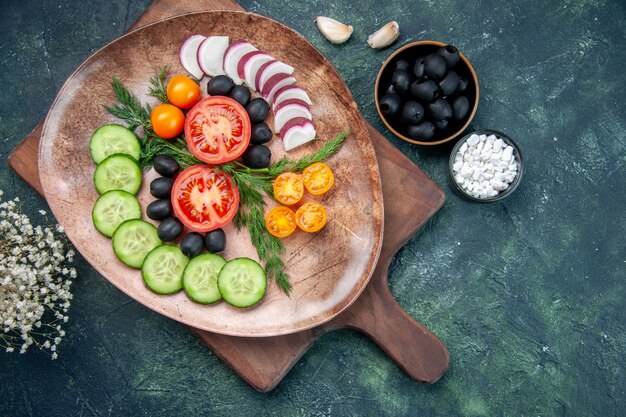 Free photo above view of fresh chopped vegetables in a brown plate on wooden cutting board olives in bowl salt garlics flower on mixed colors table