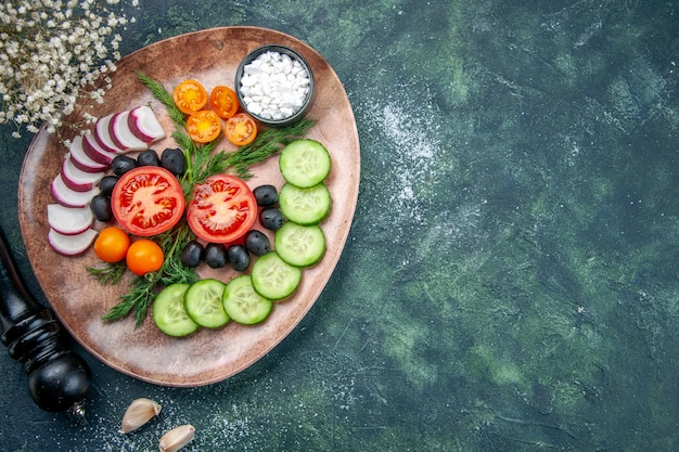 Above view of fresh chopped vegetables olives salt in a brown plate and kitchen hammer on the right side on green black mixed colors table