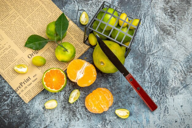 Above view of fresh citrus fruits with leaves fallen out of a black basket cut in half forms and knife on newspaper on gray background