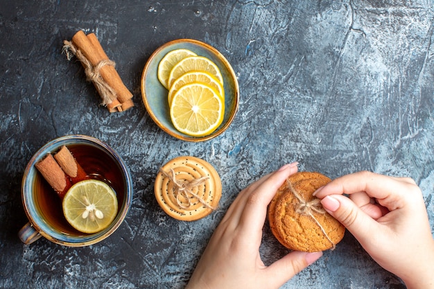 Above view of fresh lemons and a cup of black tea with cinnamon hand holding stacked cookies on dark background