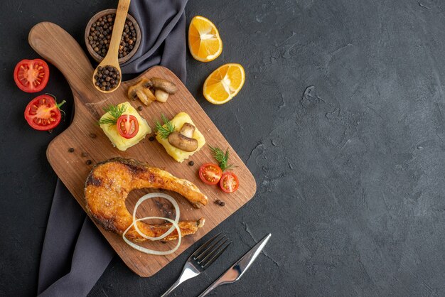 Above view of fried fish meal with mushrooms vegetables cheese on wooden board lemon slices pepper on dark color towel on the right side on black distressed surface