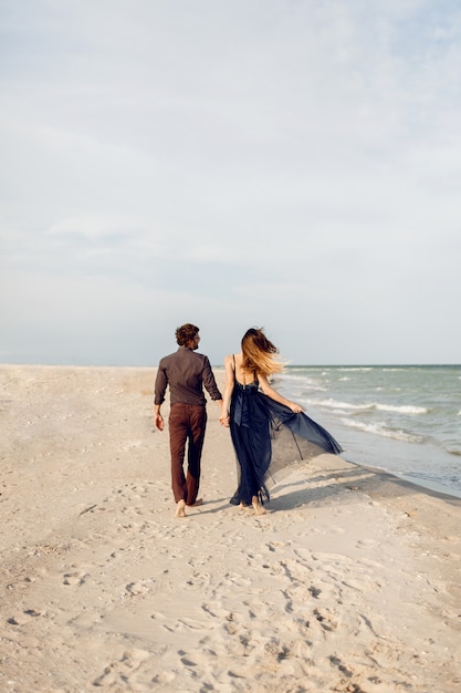 Free photo view from back. elegant couple in love walking along the beach. romantic moments. white sand and ocean waves. tropical vacation. full height.