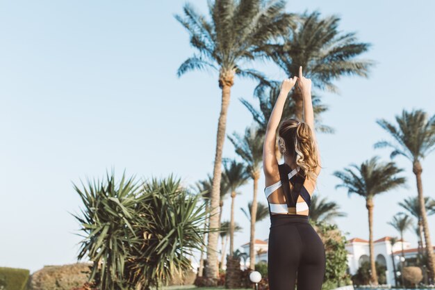 View from back joyful young woman in sportswear stretching on palm trees and blue sky. Sunny morning, expressing positivity, true emotions, healthy lifestyle.