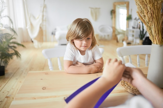 Free photo view from back of unrecognizable childâs hands holding violet paper sheet. indoor shot of cute little girl in white t-shirt sitting at desk watching her elder brother make origami. selective focus