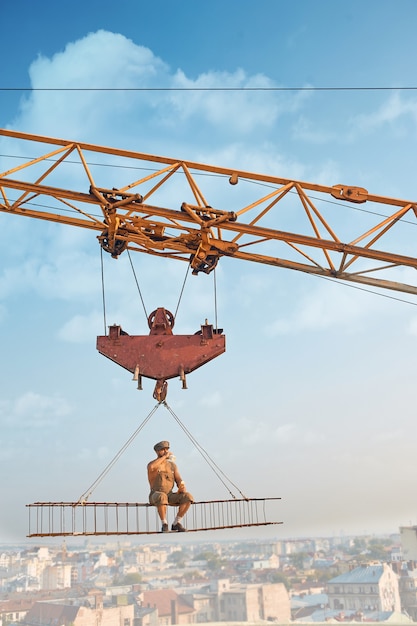 Free photo view from distance of biggest crane holding iron construction, where sitting builder and eating. man resting and looking down. cityscape on background. extreme building on high.