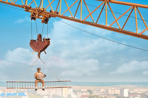 Free Photo view from distance of bold builder in work wear sitting on construction on high and reading news paper. building crane holding construction with man over city. extreme building of house in big city.