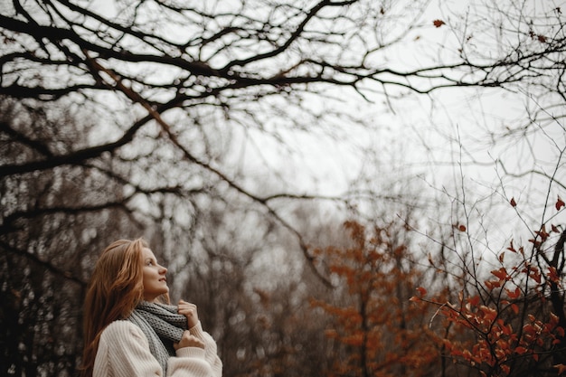 Free photo view from distance of redhead girl dressed in warm pullover standing in forest with yellow leaves