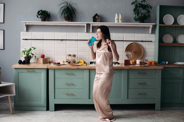 Free photo view from side of young girl wearing pajamas standing at kitchen and dancing in morning cheerful woman singing while preparing breakfast at home on weekends concept of preparation and happiness