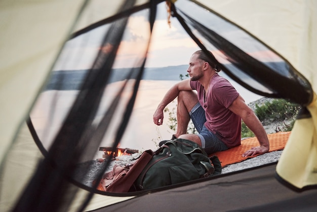 Free photo view from tent of traveller with backpack sitting on top of mountain enjoying view coast a river or lake.