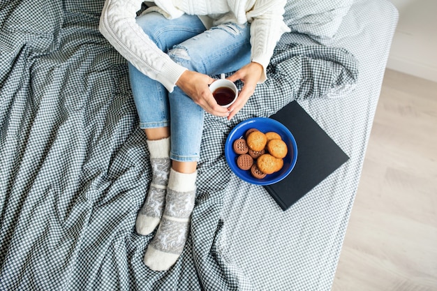 View from above of woman sitting on bed in morning, drinking coffee in cup, eating cookies, breakfast