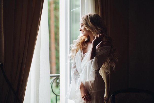 View of gorgeous woman standing near balcony and posing in white lace lingerie. Girl in vintage interior room, looking in big window. Bride at morning.