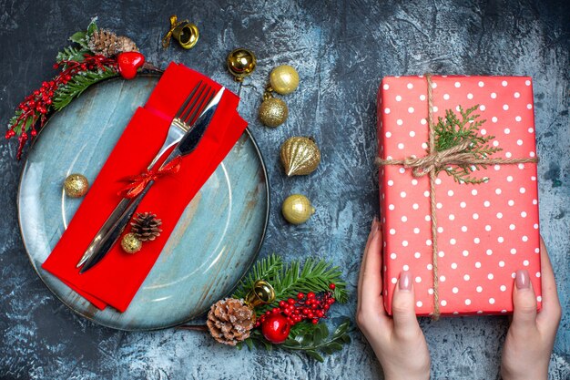 Above view of hand holding a gift box and cutlery set with red ribbon on a decorative napkin on a blue plate and christmas accessories and christmas sock on dark background