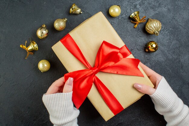 Above view of hand holding a gift box with red ribbon and decoration accessories on dark background