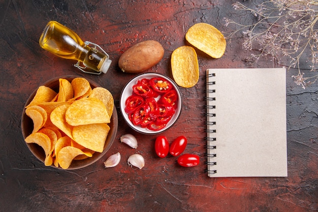 Free Photo above view of homemade delicious crispy potato chips in a brown pot fallen oil bottle ketchup tomatoes potato garlic and notebook on dark background