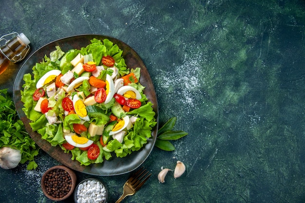 Above view of homemade delicious salad with many ingredients in a plate and spices fallen oil bottle fork on black green mix colors table with free space