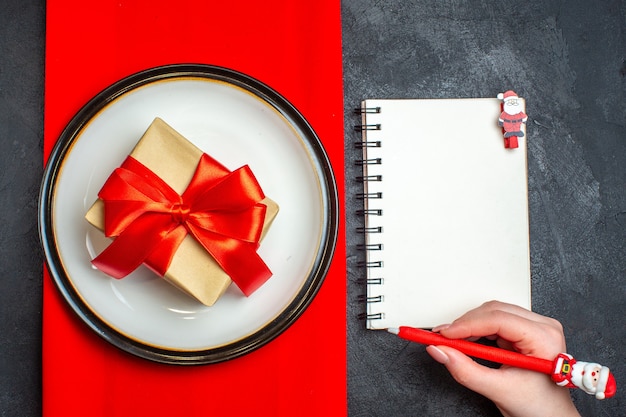 Free photo above view of national christmal meal background with gift with bow-shaped red ribbon on empty plates on a red napkin and hand holding a pen on notebook on black background