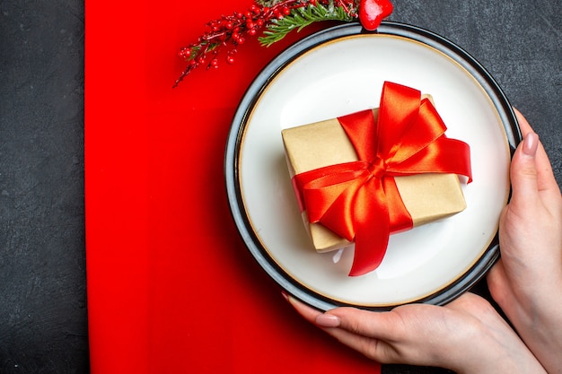 Above view of national Christmal meal background with hand holding empty plates with bow-shaped red ribbon and fir branches on a red napkin on black table