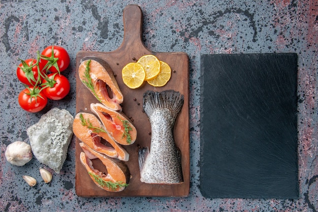 Above view of raw fishes lemon slices greens pepper on wooden cutting board and vegetables cutlery set on dark color tray on blue black colors table