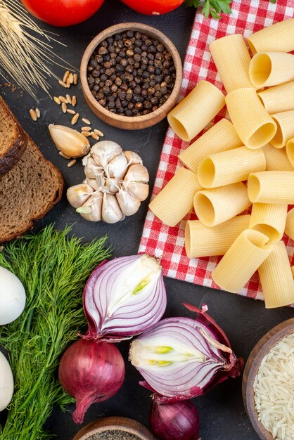 Above view of raw pastas on red stripped towel rice in a brown bowl fresh vegetables bread slices on black table