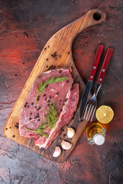 Above view of red meat on wooden cutting board and garlic green pepper oil bottle fork and knife on dark background