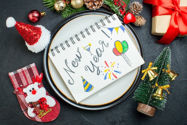 Above view of spiral notebook with pen on dinner plate christmas tree fir branches conifer cone gift box santa claus hat christmas sock on black background