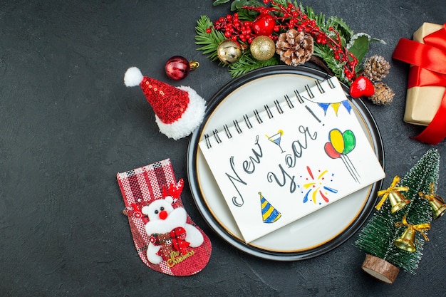 Above view of spiral notebook with pen on dinner plate christmas tree fir branches conifer cone gift box santa claus hat christmas sock on the left side on black background